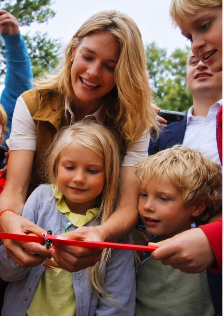Une femme blonde et des enfants coupant un ruban rouge lors d'une inauguration, en collaboration avec Naked Heart Foundation.