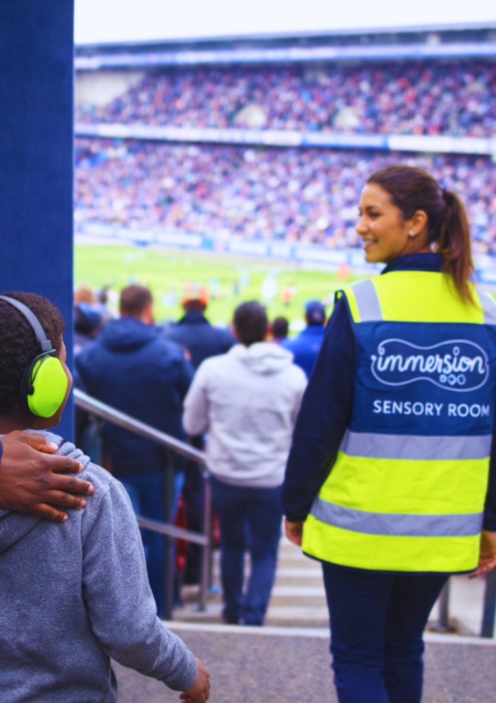 Une accompagnatrice de Big Impacts portant un gilet "Sensory Room" guide un enfant avec un casque anti-bruit vers les tribunes d'un stade.